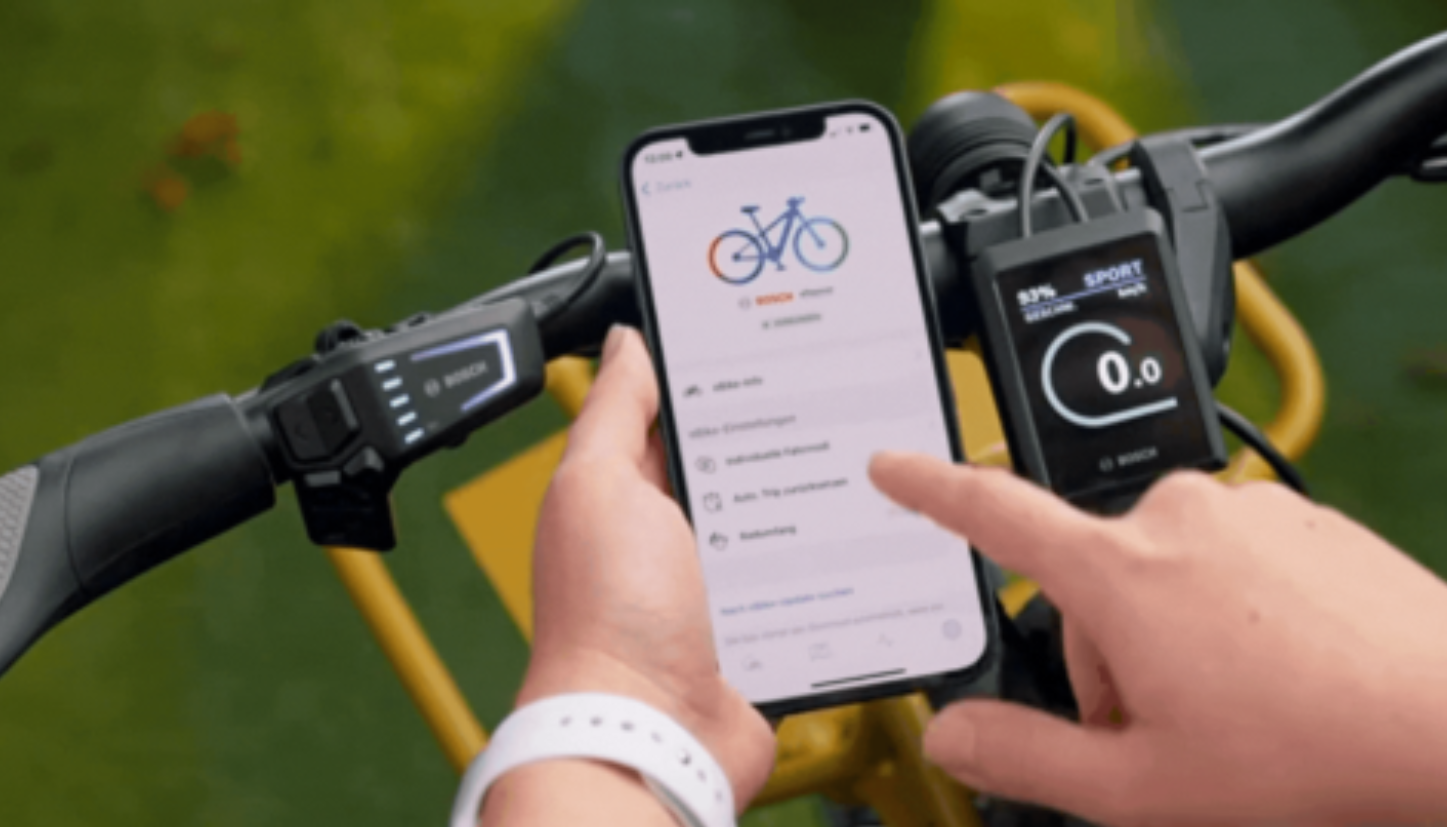 Afbeelding van een telefoon met de Bosch E-bike Flow App. Met een elektrische fiets met telefoonhouder op de achtergrond.